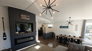 A modern living room with a flat-screen TV, a fireplace, a dining area with a long table, and a kitchen in the background. - Turtle Loft Lakefront Cabin Townhome (Kootenay Boundary E / West Bo*)