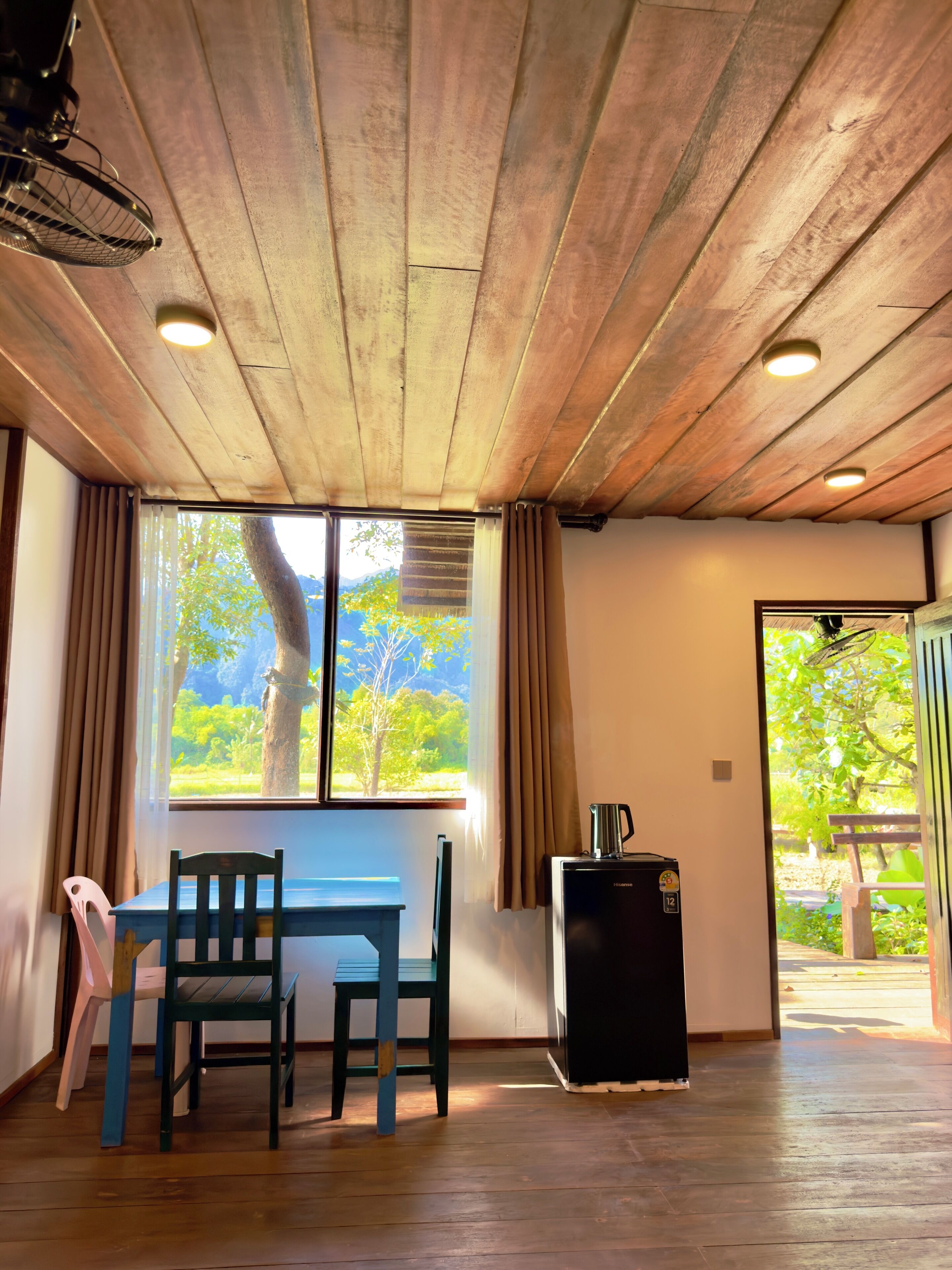 A room with wooden ceiling, a table with chairs, a small black refrigerator, and a window with curtains.