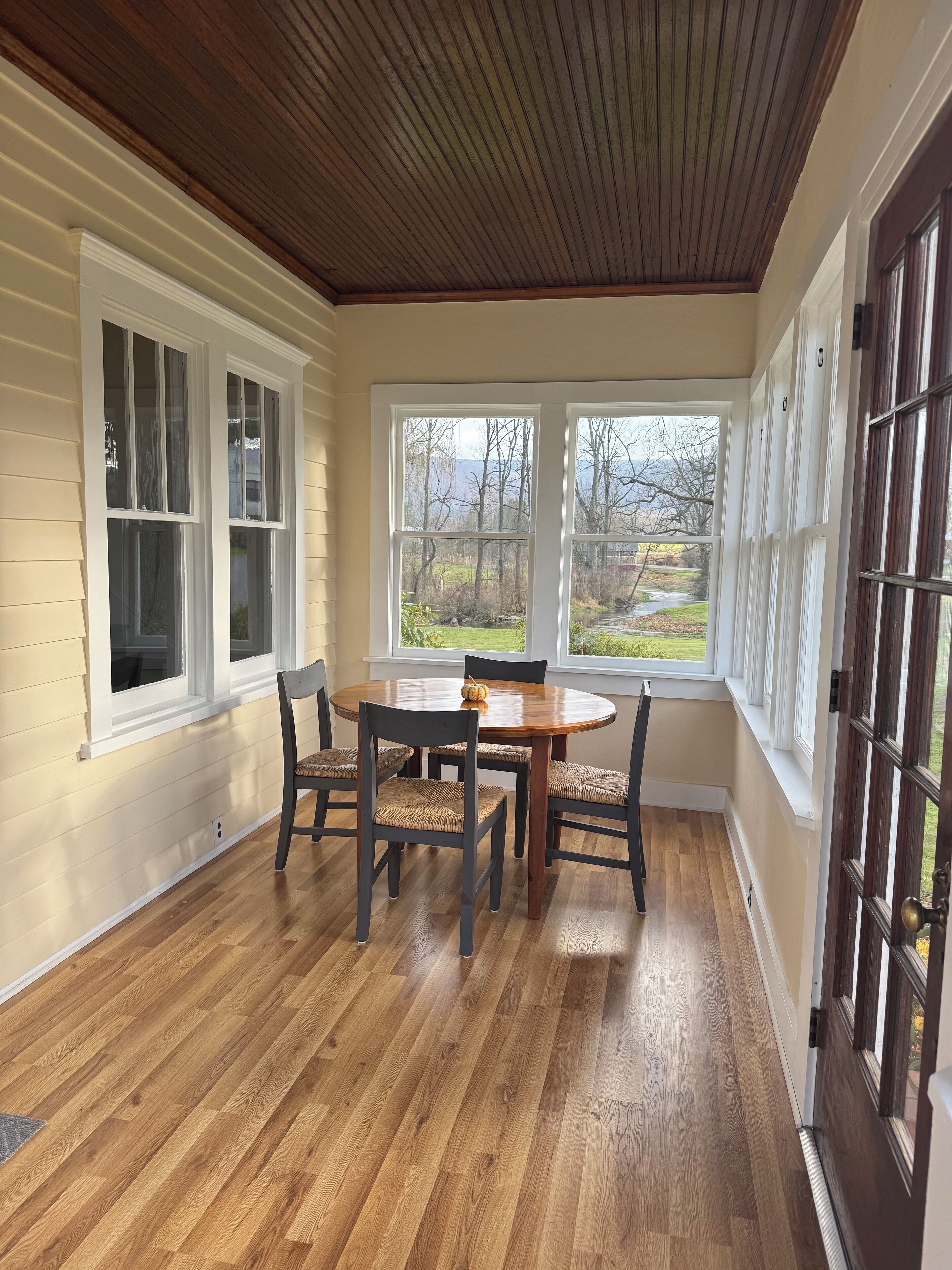 A dining area with a wooden table and chairs, large windows, and a view of a landscape.