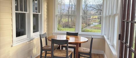 A dining area with a wooden table and chairs, large windows, and a view of a landscape.