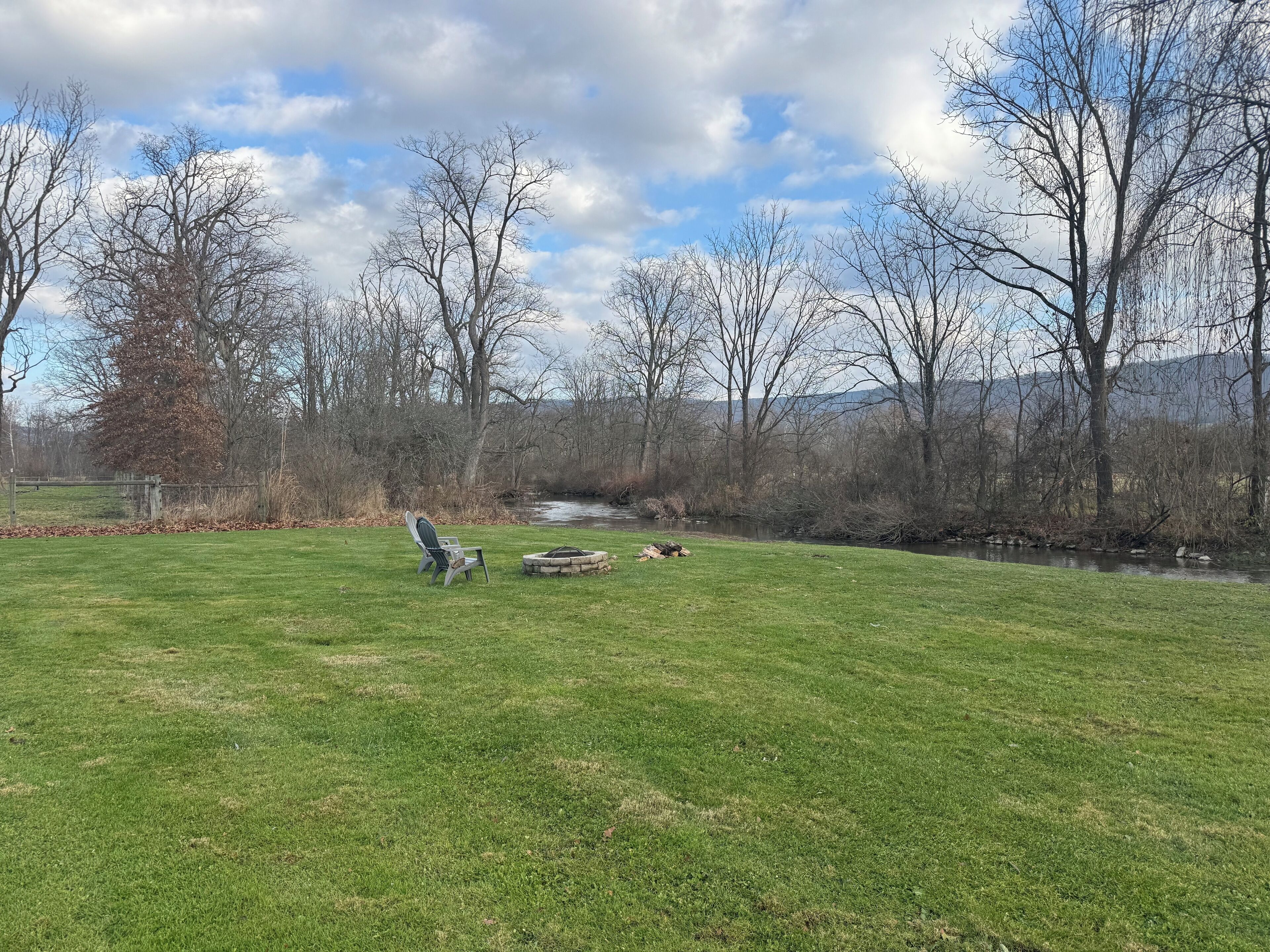 A grassy field with a chair and a fire pit, surrounded by trees and a river.