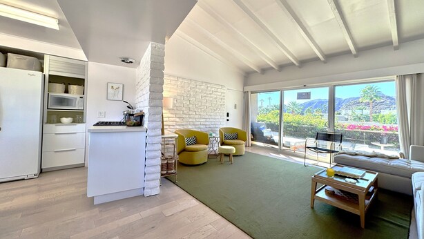 A modern kitchen with white cabinetry and a built-in microwave. There is a dining area with yellow chairs and a green rug. A large window offers a view of the outdoors. - Desert Retreat in Rancho Mirage – Bright, Stylish Condo with Resort Amenities (Rancho Mirage)