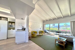 A modern kitchen with white cabinetry and a built-in microwave. There is a dining area with yellow chairs and a green rug. A large window offers a view of the outdoors. - Desert Retreat in Rancho Mirage – Bright, Stylish Condo with Resort Amenities (Rancho Mirage)