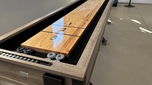 Une table de billard avec un plateau en bois et un drap noir, située dans une pièce moderne dotée d’une fenêtre et d’un coin bar.