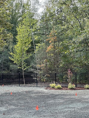 A gravel area with a black metal fence, surrounded by trees with green and autumn-colored leaves. - SOMETHING FOR EVERYONE,  New 3+ BR, 4 Bath, Ranch, Game Rm, Fenced Yd & Pond (Fennville)