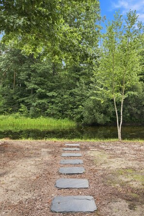 A stone pathway leading through a natural area with dense greenery and a body of water. - SOMETHING FOR EVERYONE,  New 3+ BR, 4 Bath, Ranch, Game Rm, Fenced Yd & Pond (Fennville)
