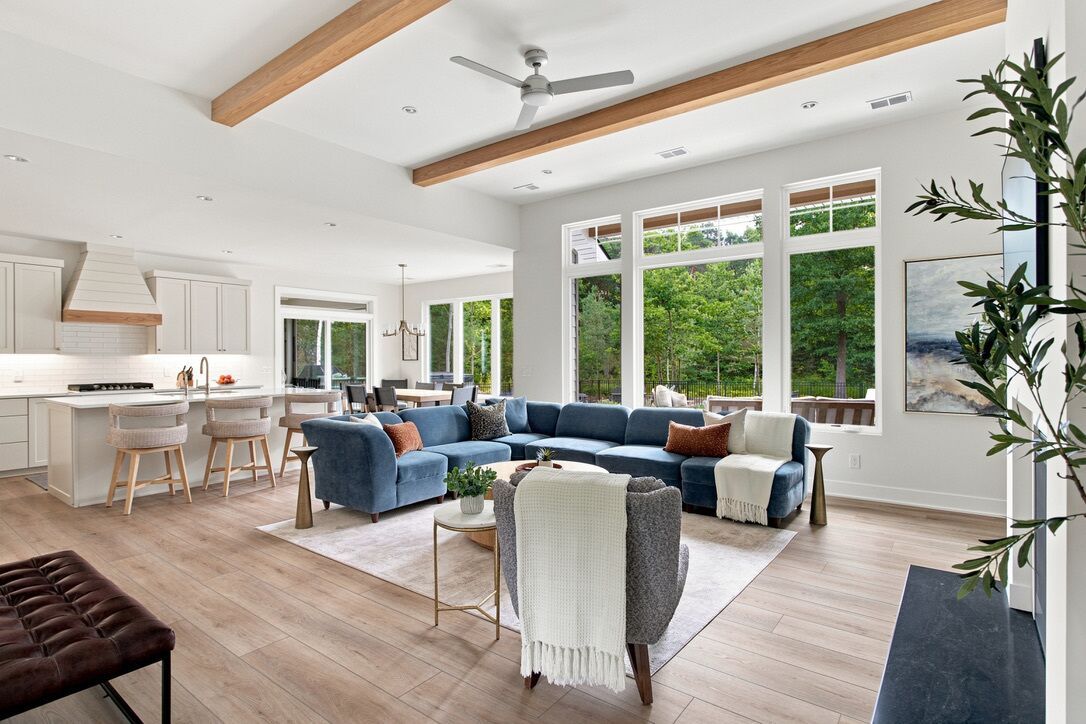 A spacious living area with a blue sectional sofa, a coffee table, and a fireplace. There is a kitchen area with white cabinets and a dining table. The room has large windows offering a view of greenery outside.