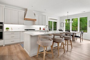 A modern kitchen with white cabinetry, a central island with bar stools, and a large window offering a view of greenery. - SOMETHING FOR EVERYONE,  New 3+ BR, 4 Bath, Ranch, Game Rm, Fenced Yd & Pond (Fennville)