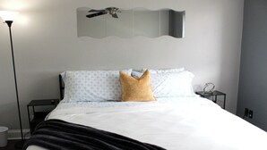 Ein ordentlich bezogenes Bett mit einem gemusterten Kopfkissen, einer schwarzen Tagesdecke und einer Wandleuchte.