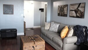 Ein Wohnzimmer mit grauem Sofa, einem Korbwäscheschrank und zwei an der Wand hängenden Bildern.