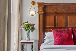 Een slaapkamer met een houten hoofdbord, een nachtkastje met een bloemenvaas, en een raam met gordijnen.