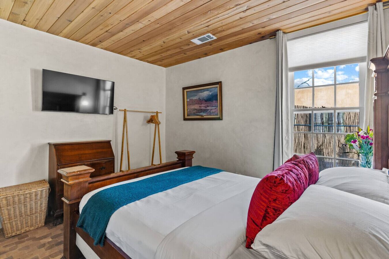 Une chambre avec un plafond en bois, un lit avec une literie blanche et rouge, un téléviseur fixé au mur, un tableau et une fenêtre donnant sur un paysage.