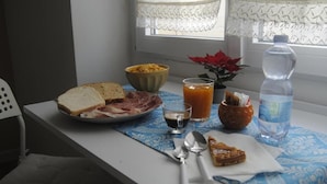 A breakfast table with bread, meat, a bowl of cereal, a glass of orange juice, a bottle of water, and a spoon. - Coro & Bentu  (Alghero)