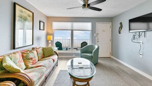 Ein Wohnzimmer mit einer Couch, einem Glastisch, einem Deckenventilator, einem Balkon mit Sitzgelegenheiten und einem an der Wand befestigten Fernseher.