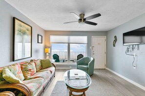 A living room with a sofa, a glass-top coffee table, a ceiling fan, a balcony with chairs, and a TV mounted on the wall. - Charming 2-bedroom condo in beautiful Galveston with Ocean view (Galveston)