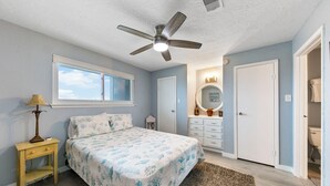 Ein Schlafzimmer mit einem Bett, einem Deckenventilator, einem Fenster, einem Schminktisch und einem durch eine offene Tür einsehbaren Badezimmer.