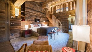 Un dormitorio rústico con paredes de madera, cama, mesitas de noche, una silla, una pequeña mesa y una escalera.