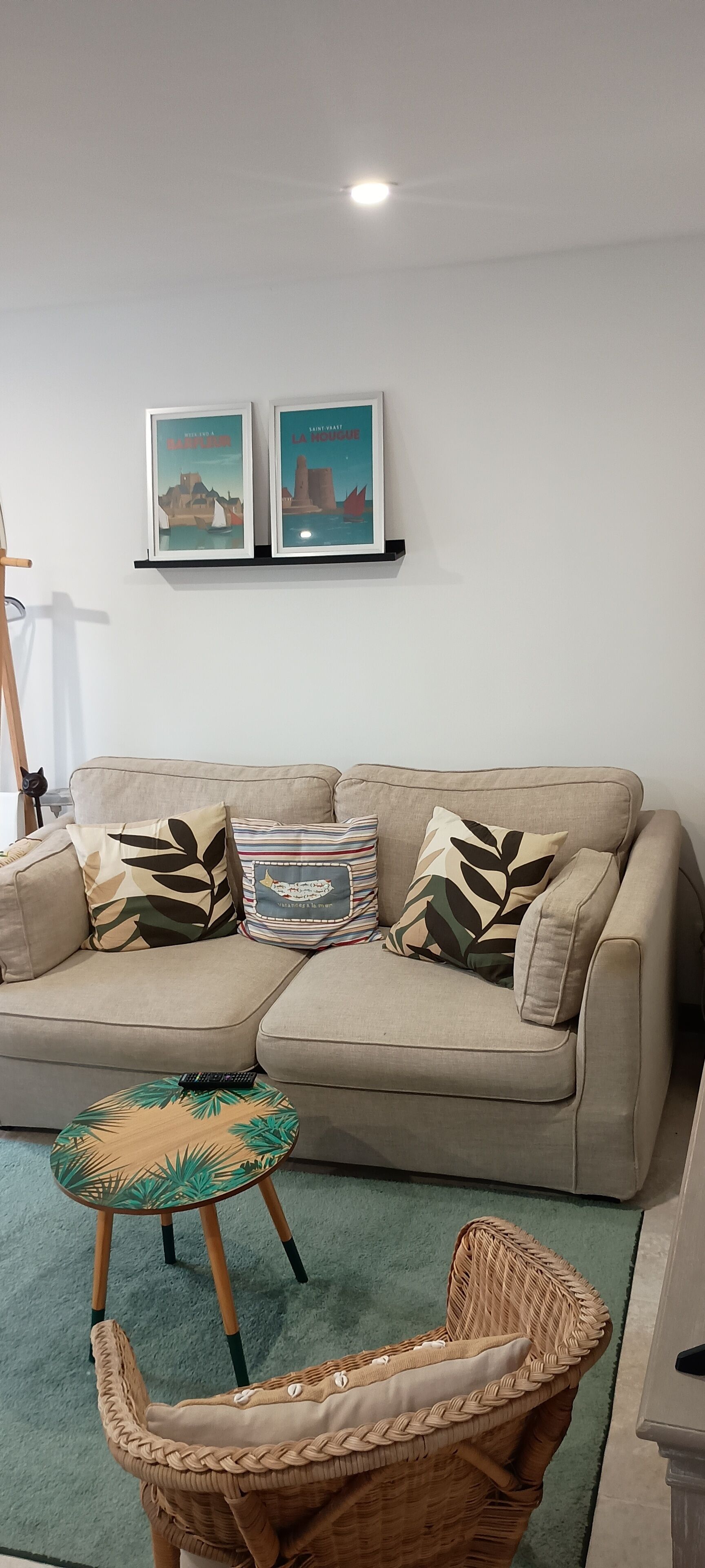 Ein Wohnzimmer mit einem beigefarbenen Sofa, zwei gerahmten Bildern an der Wand, einem kleinen runden Tisch und einem Korbstuhl.