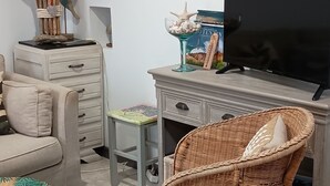 Una sala de estar con sofá, una silla de mimbre, una mesa de comedor, televisor y un cuadro enmarcado en la pared.