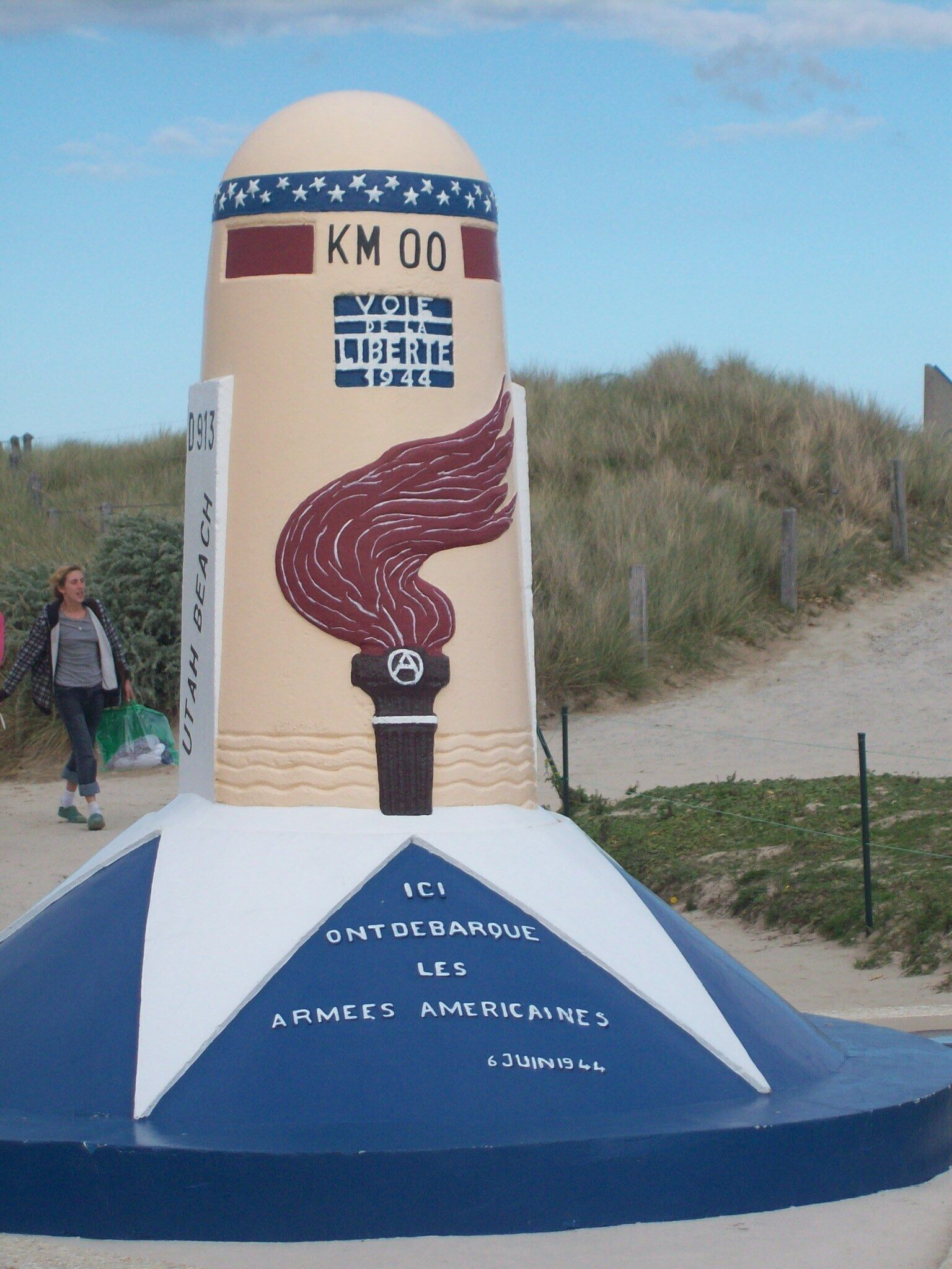 Ein Denkmal mit einem Sternmuster, einer geflügelten Fackel und Inschriften, darunter "KM 00", "VOI DE LA LIBERTE 1944" und "ICI ONT DEBARQUE LES ARMEES AMERICAINES 6 JUIN 1944".