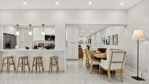 A modern kitchen with white cabinetry, a central island with stools, and a dining area with a wooden table and chairs. - Delray Oasis: Heated Pool, Spa, Arcade, Sleeps 10 (Delray Beach)