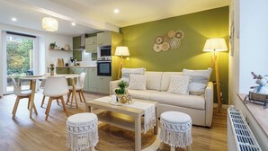 A modern living room with a sofa, coffee table, and round ottomans. There is a dining area with chairs and a table. The room has a green accent wall and a large window. - Cosy house with terrace in the heart of Munster (Munster)