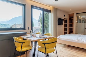 Ein modernes Schlafzimmer mit einem hölzernen Bett, einem Essbereich mit gelben Stühlen, einem großen Fenster mit Blick auf Berge und einem an der Wand montierten Fernseher.