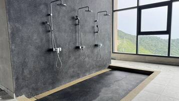 A modern bathroom with three wall-mounted showers, a large window with a view of greenery, and a tiled floor.