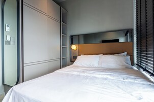 Una cama bien hecha con sábanas blancas, una lámpara de noche y un amplio armario con espejo.