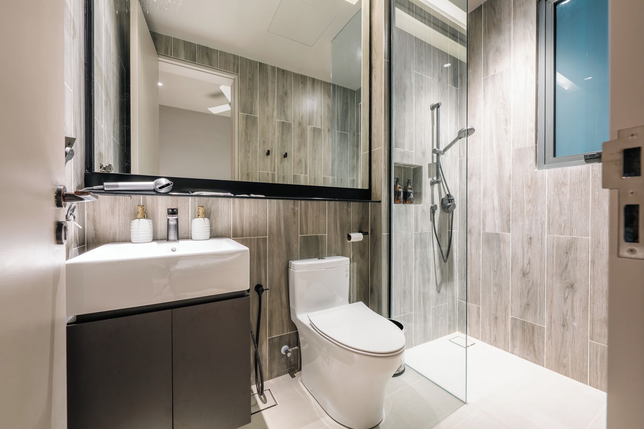 A modern bathroom with a white sink, toilet, and shower. It features a large mirror, wooden wall tiles, and a glass shower enclosure.