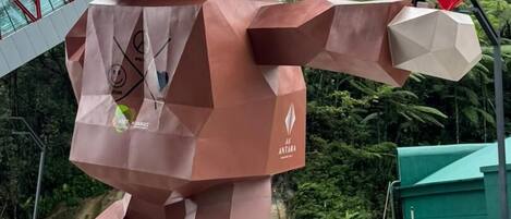 A large, geometric sculpture resembling a figure with a heart in its hand, located in an urban setting with a modern building in the background.