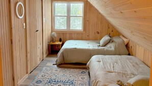 Un dormitorio acogedor en el ático con paredes de madera, un ventanal grande, dos camas, una mesita de noche y una alfombra estampada.