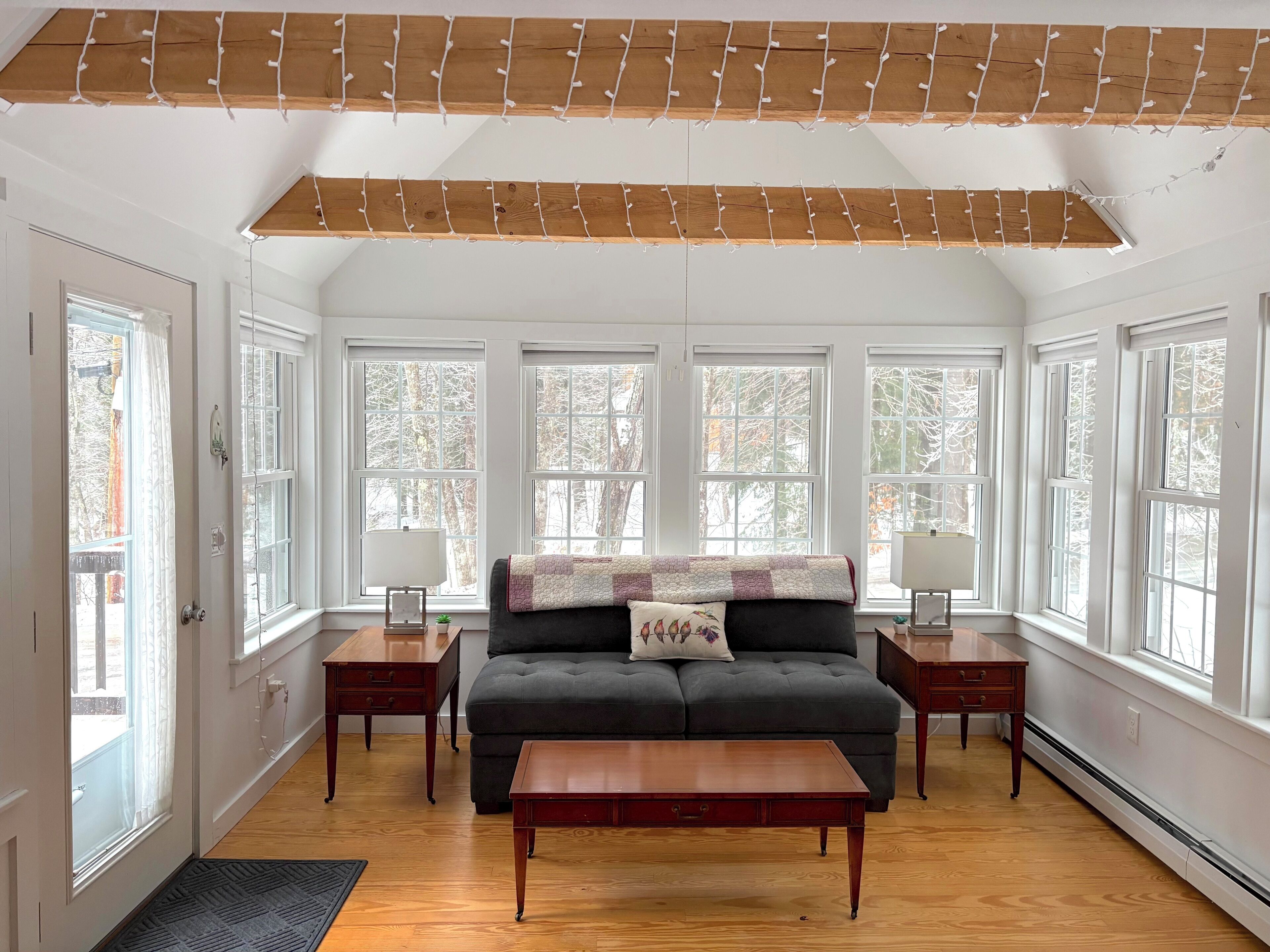 Un salón amplio con un sofá oscuro, una mesa de centro de madera y dos mesitas auxiliares. Ventanas grandes ofrecen luz natural y vistas a árboles.