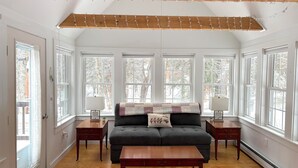 Un salón amplio con un sofá oscuro, una mesa de centro de madera y dos mesitas auxiliares. Ventanas grandes ofrecen luz natural y vistas a árboles.