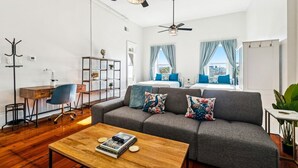 Un salon moderne avec un canapé gris, une table basse en bois et un bureau avec une chaise. Il y a deux fenêtres avec des rideaux, un ventilateur de plafond et une plante sur la droite.