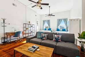 Un salon moderne avec un canapé gris, une table basse en bois et un bureau avec une chaise. Il y a deux fenêtres avec des rideaux, un ventilateur de plafond et une plante sur la droite.