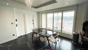 Un espace de salle à manger moderne avec une table et des chaises en verre, un lustre et une grande fenêtre donnant sur la ville.