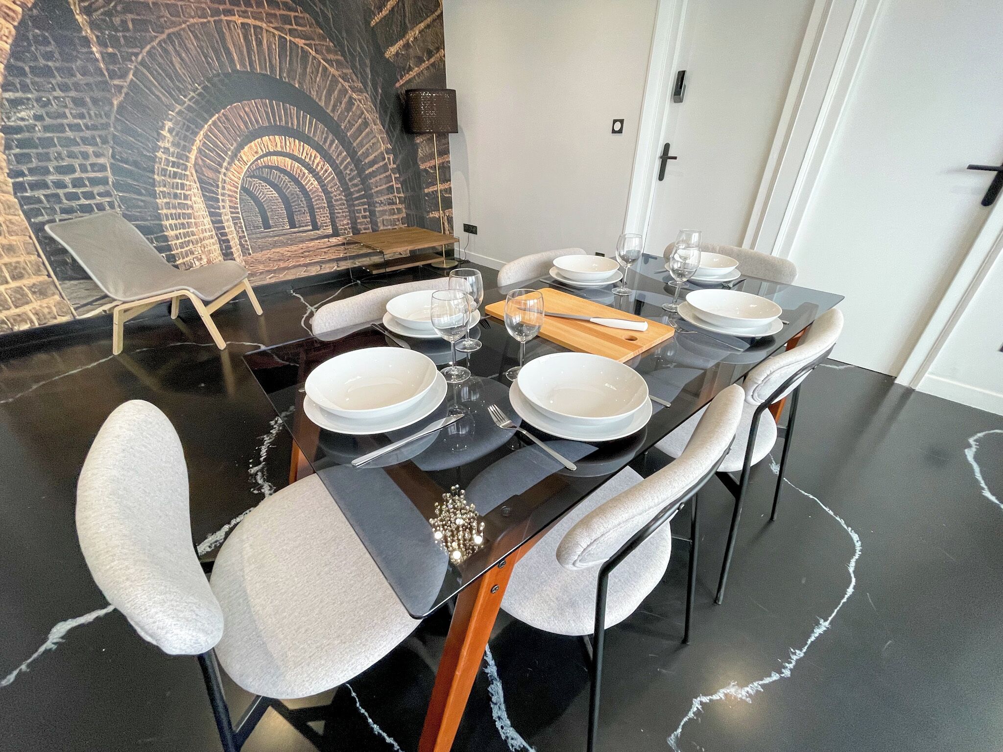 A dining area with a black table set for six, white plates, and cutlery. There is a wall mural of an arched tunnel and a grey lounge chair in the background.