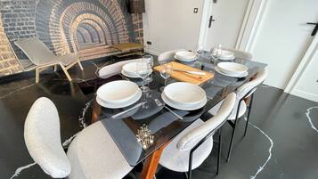 A dining area with a black table set for six, white plates, and cutlery. There is a wall mural of an arched tunnel and a grey lounge chair in the background.