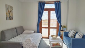 Et soveværelse med en seng, en grå sofa, et lille bord med en skål og bøger, samt et vindue med blå gardiner.