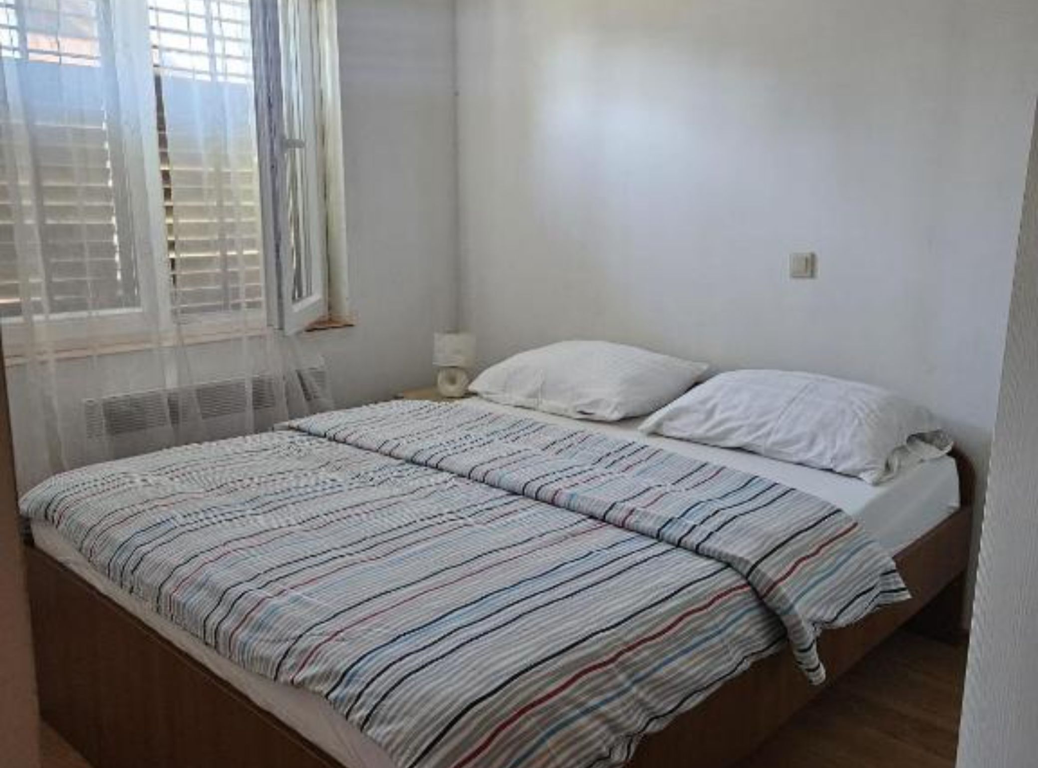 Quarto com cama, paredes brancas, janela com persianas e mesa de cabeceira com abajur.
