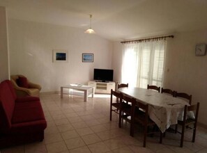 Un soggiorno con un divano rosso, un tavolo da pranzo con sedie, una TV su un mobile basso e una finestra con tende.