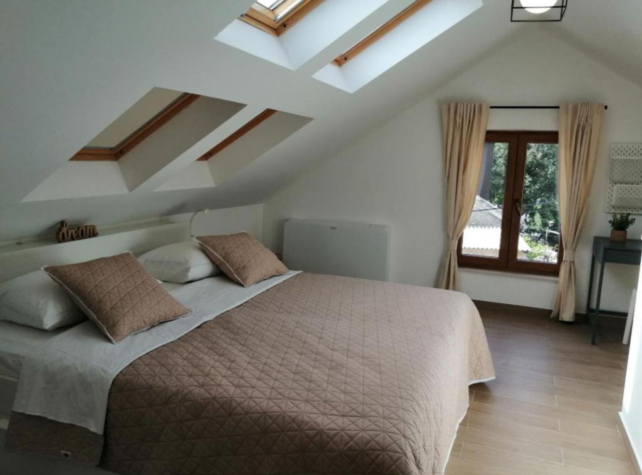 A bedroom with a sloped ceiling, a bed with a quilted bedspread, two pillows, a bedside table with a lamp, and a window with curtains.