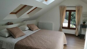 A bedroom with a sloped ceiling, a bed with a quilted bedspread, two pillows, a bedside table with a lamp, and a window with curtains. - House Serenity (Vrgorac)