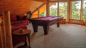 Una habitación acogedora con mesa de billar, un sofá rojo y amplios ventanales con vistas a árboles.