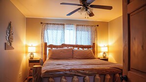 Un dormitorio con estructura de cama de madera, dos mesitas de noche con lámparas, un ventilador de techo y una ventana con cortinas estampadas.