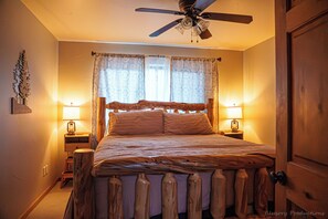 Ein Schlafzimmer mit einem hölzernen Bettgestell, zwei Nachttischen mit Lampen, einem Deckenventilator und einem Fenster mit gemusterten Vorhängen.
