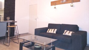 A modern living room with a glass coffee table, a dark sofa with two pillows, and a kitchen area with a bar stool and wine glasses. - Comfortable 3-bedroom T4 apartment (Mourenx)
