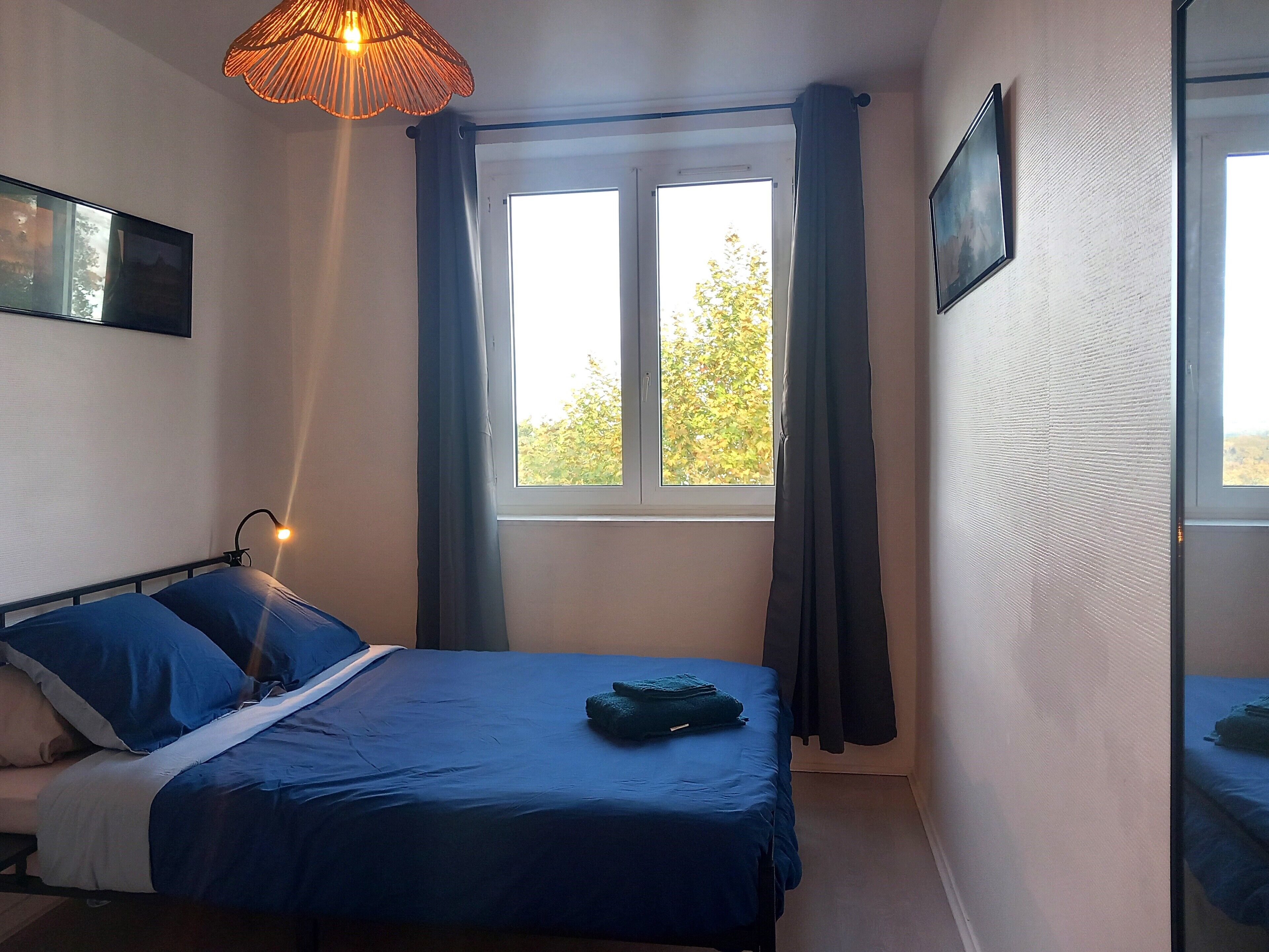 Un dormitorio con cama, lámpara de noche, ventana con cortinas y televisión instalada en la pared.