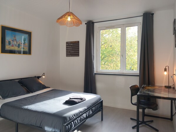 A bedroom with a bed, a desk, a chair, a hanging light fixture, a framed picture, and a departure board.